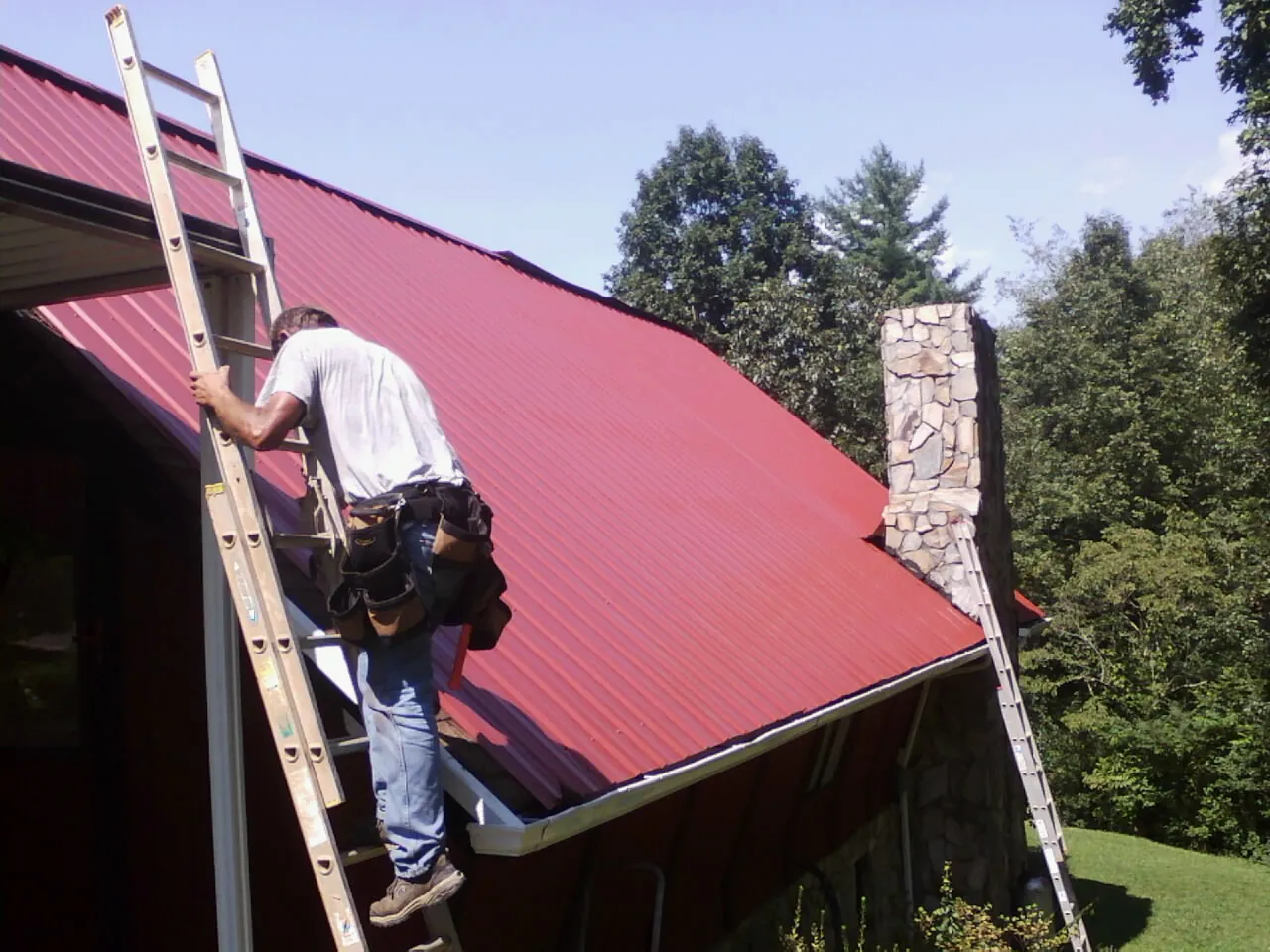 Roofer on a ladder beside a red metal roof and stone chimney — Soffit & Fascia Repair by Roofing Brothers in Garden Grove